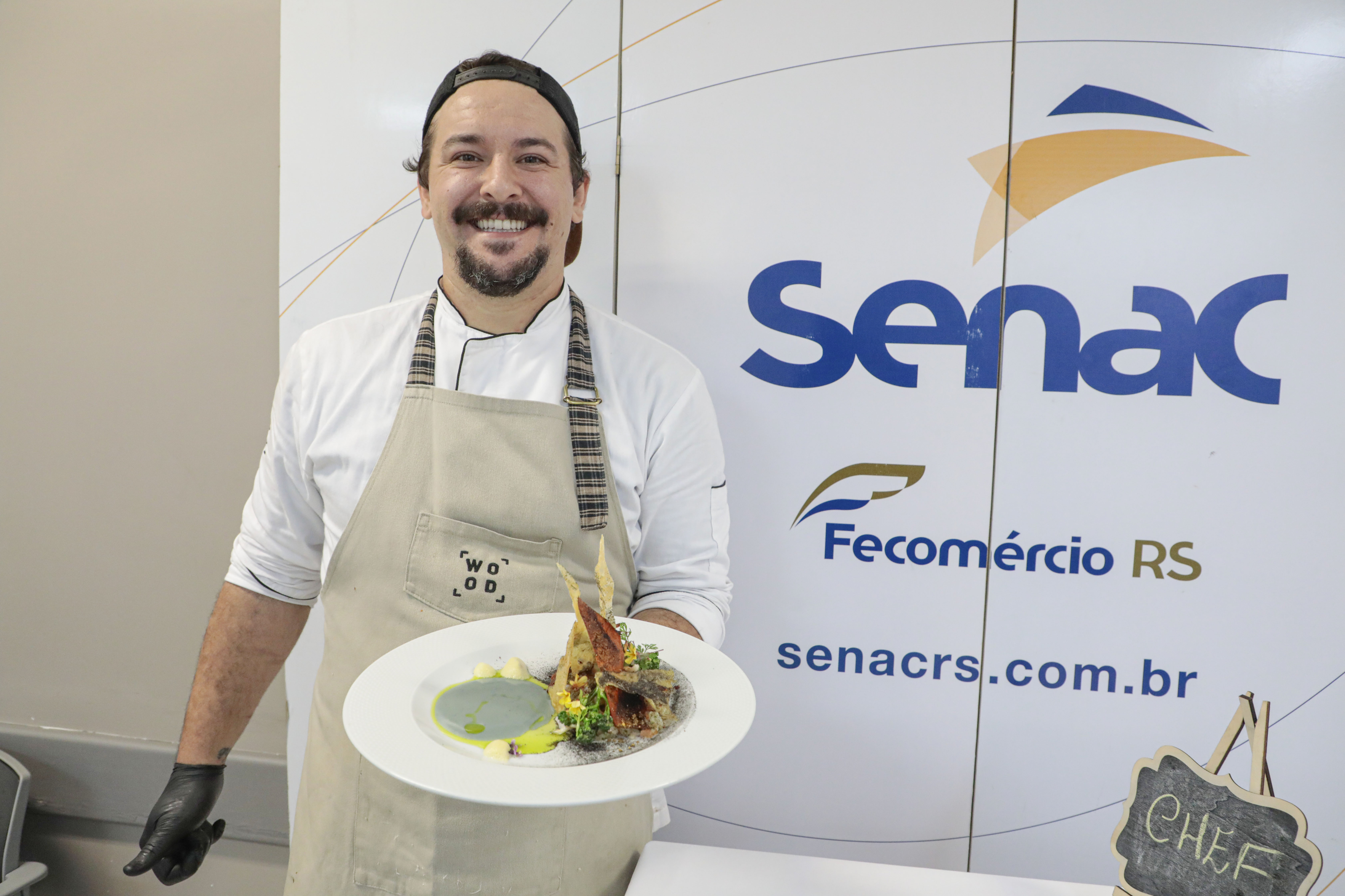 Chef sorri para a foto com um prato na mão. Ele está vestindo um avental e ao fundo aparece um banner escrito Senac.