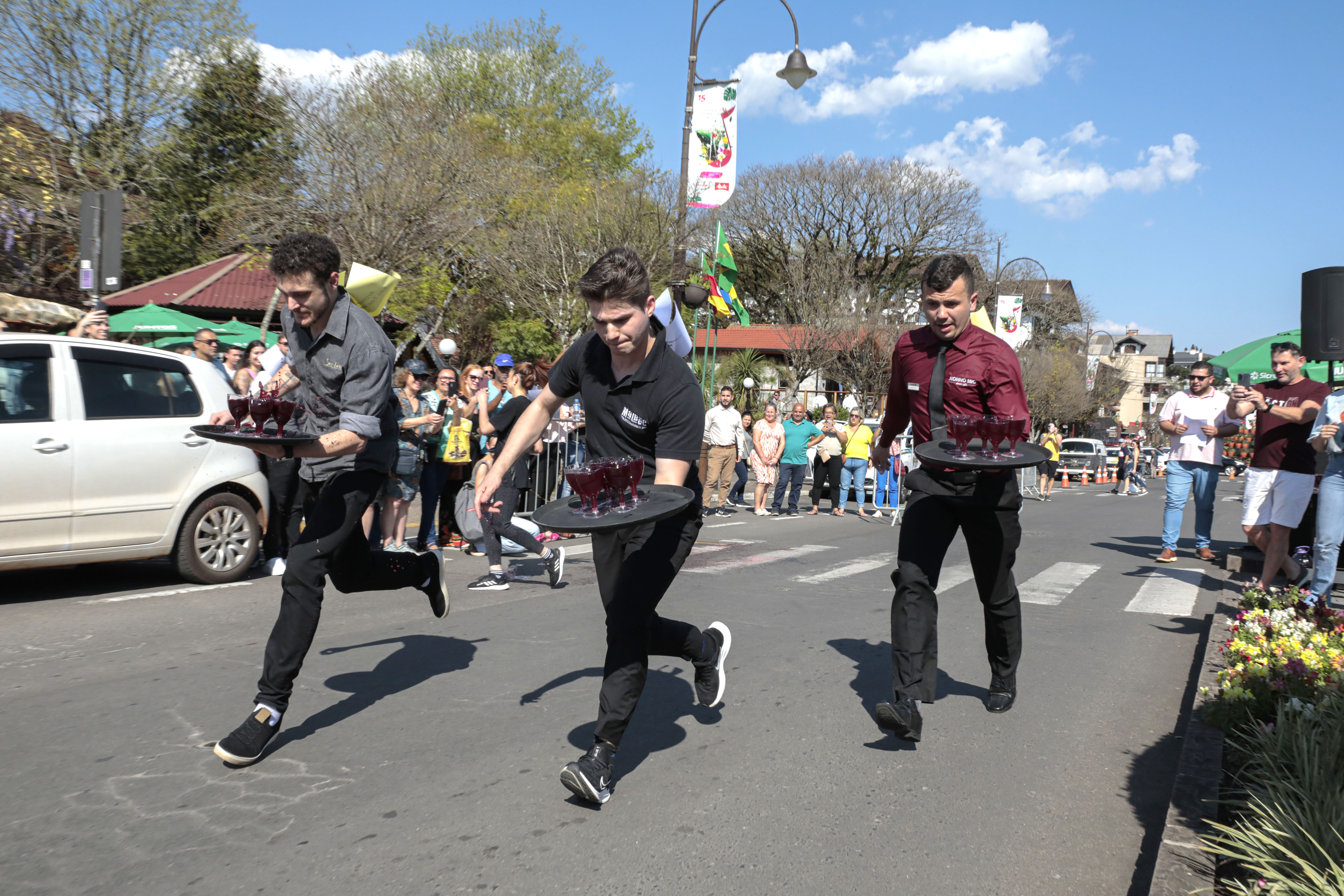 Garçons estão correndo e carregando bandejas com taças cheias em participação na Corrida de Garçons do Festival de Cultura e Gastronomia de Gramado.